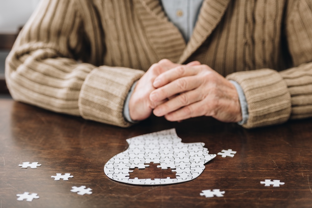 Person in beige cardigan with clasped hands sitting at wooden table with incomplete white puzzle pieces arranged in shape of human head, representing mental health, memory, or cognitive challenges with scattered puzzle pieces on table surface