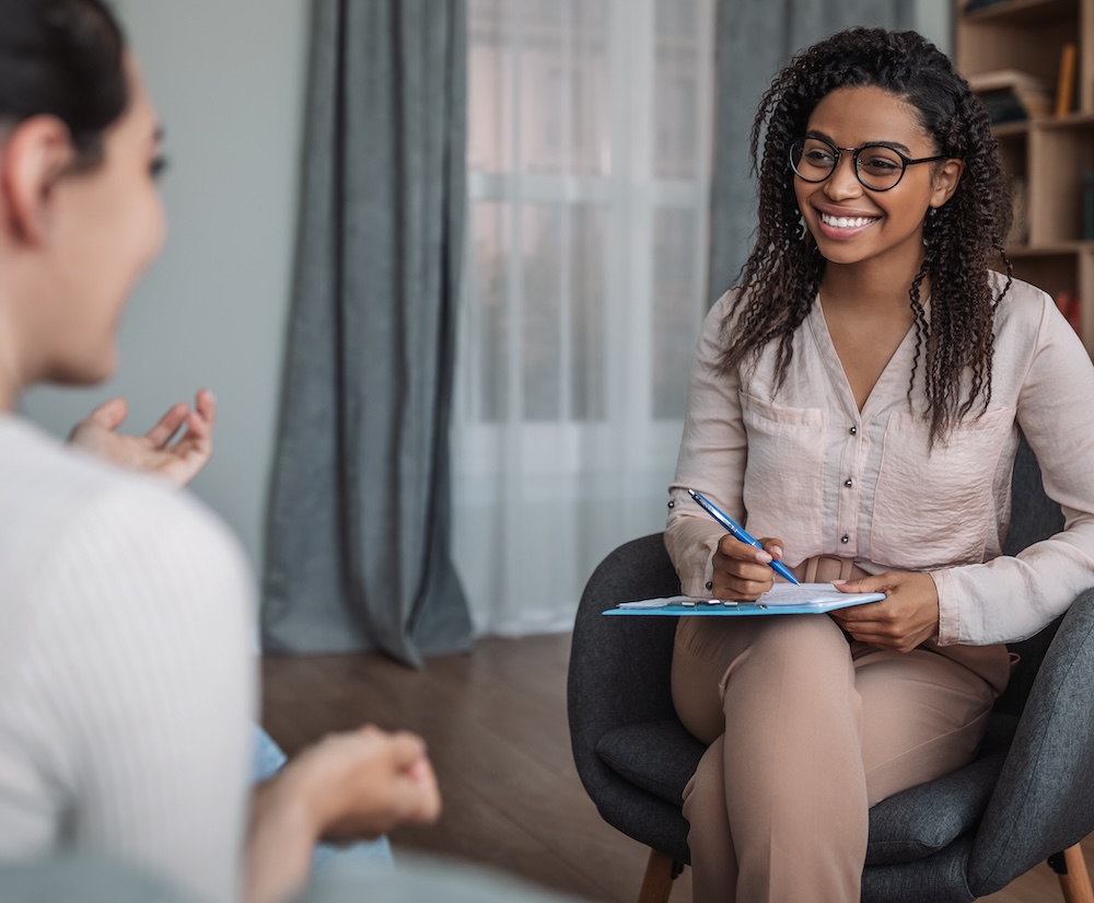Clinical psychologist with clipboard smiling during therapy session with client in comfortable office setting representing compassionate mental health care