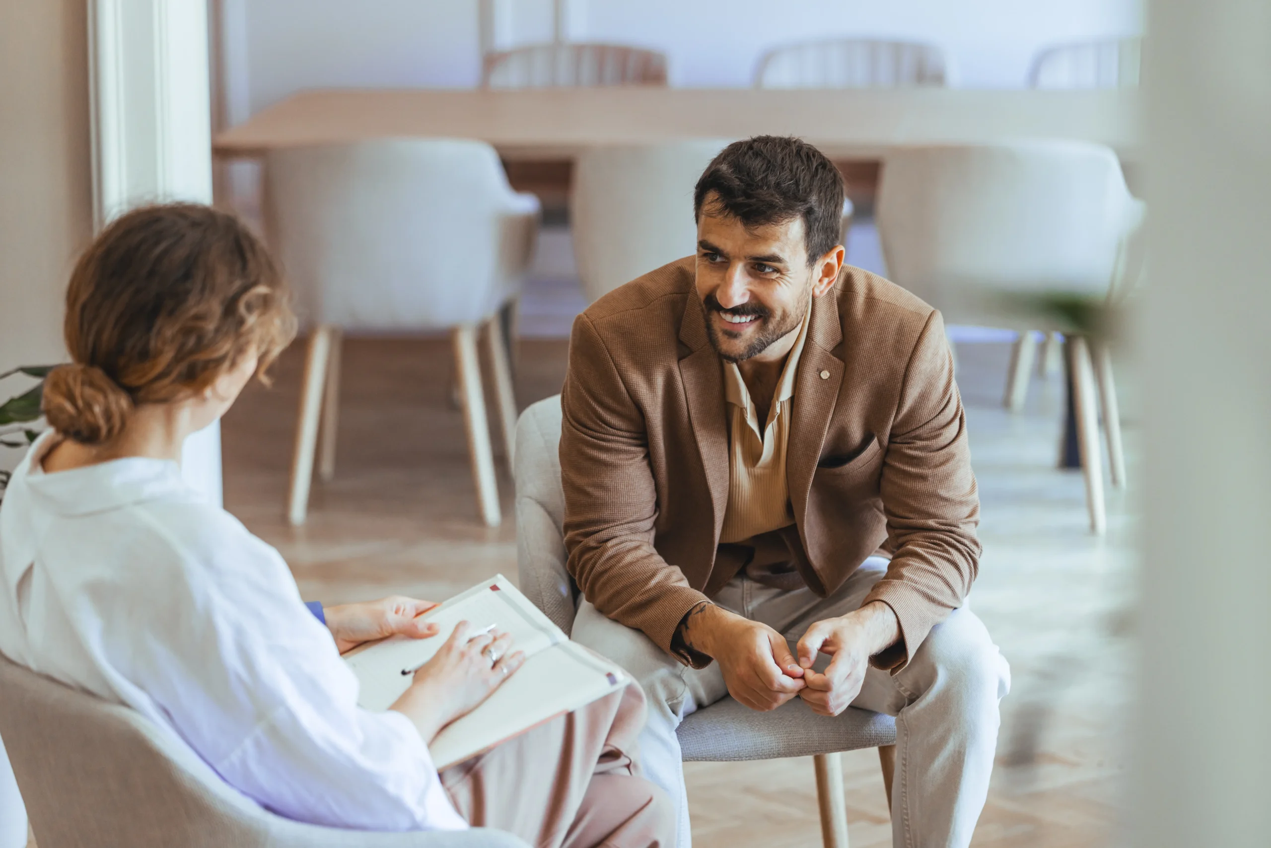 Man in brown blazer smiling while speaking with therapist during counseling session, therapist holding notepad in bright, modern office with minimalist furniture