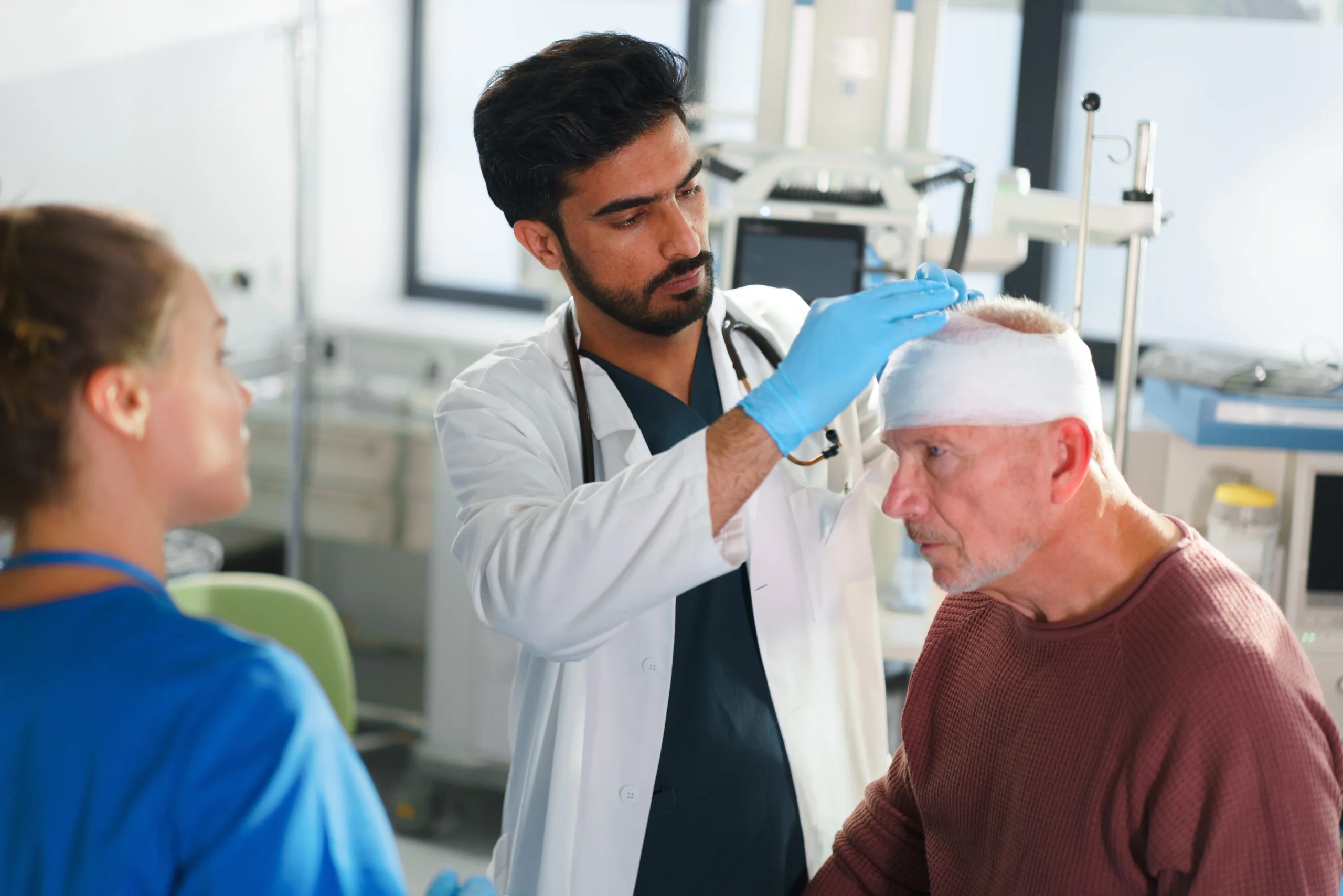 Male doctor with stethoscope examining senior patient with head bandage while female nurse in blue scrubs observes in modern medical clinic