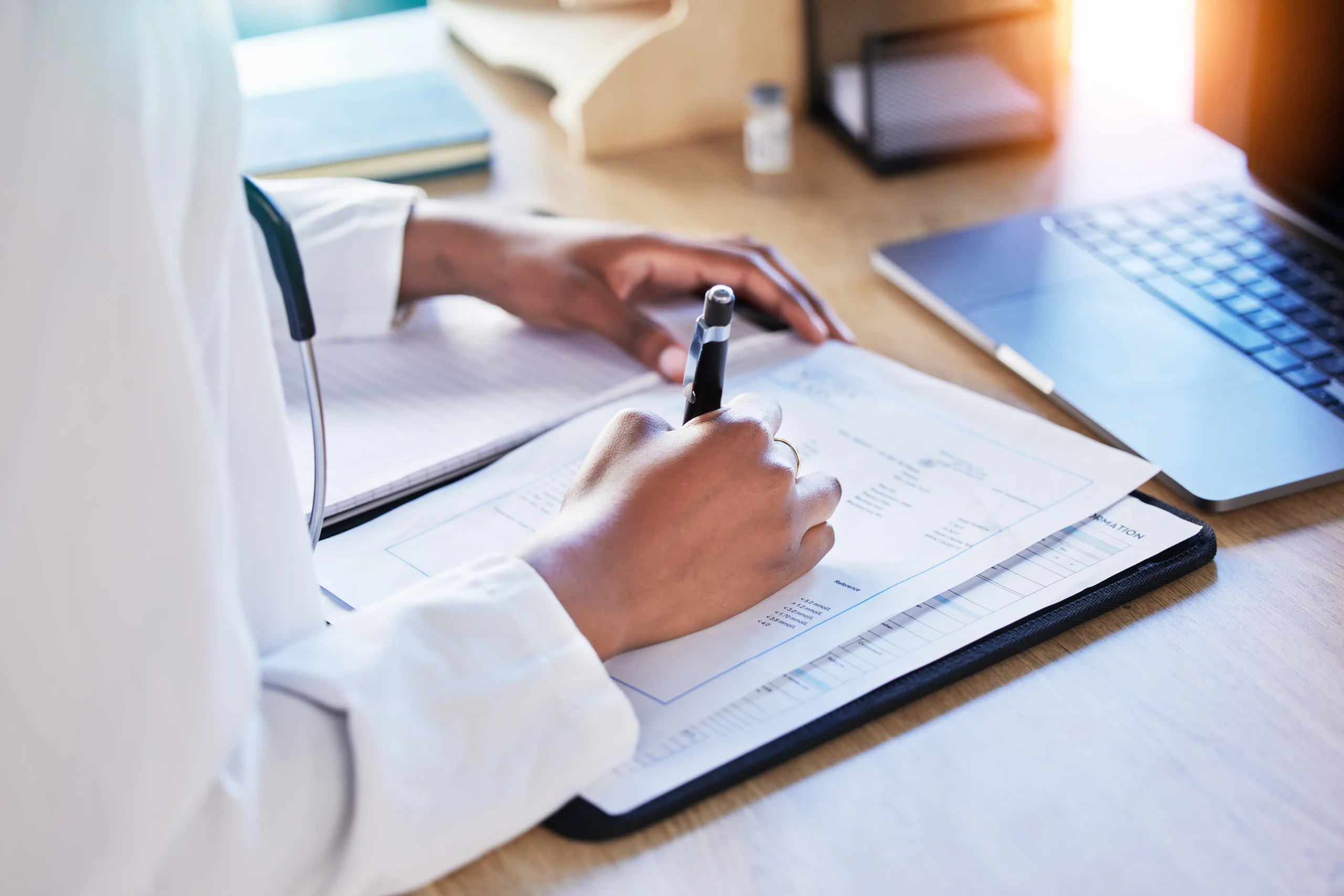 Healthcare professional in white coat writing on medical documents attached to a clipboard at a wooden desk with laptop keyboard visible in the background