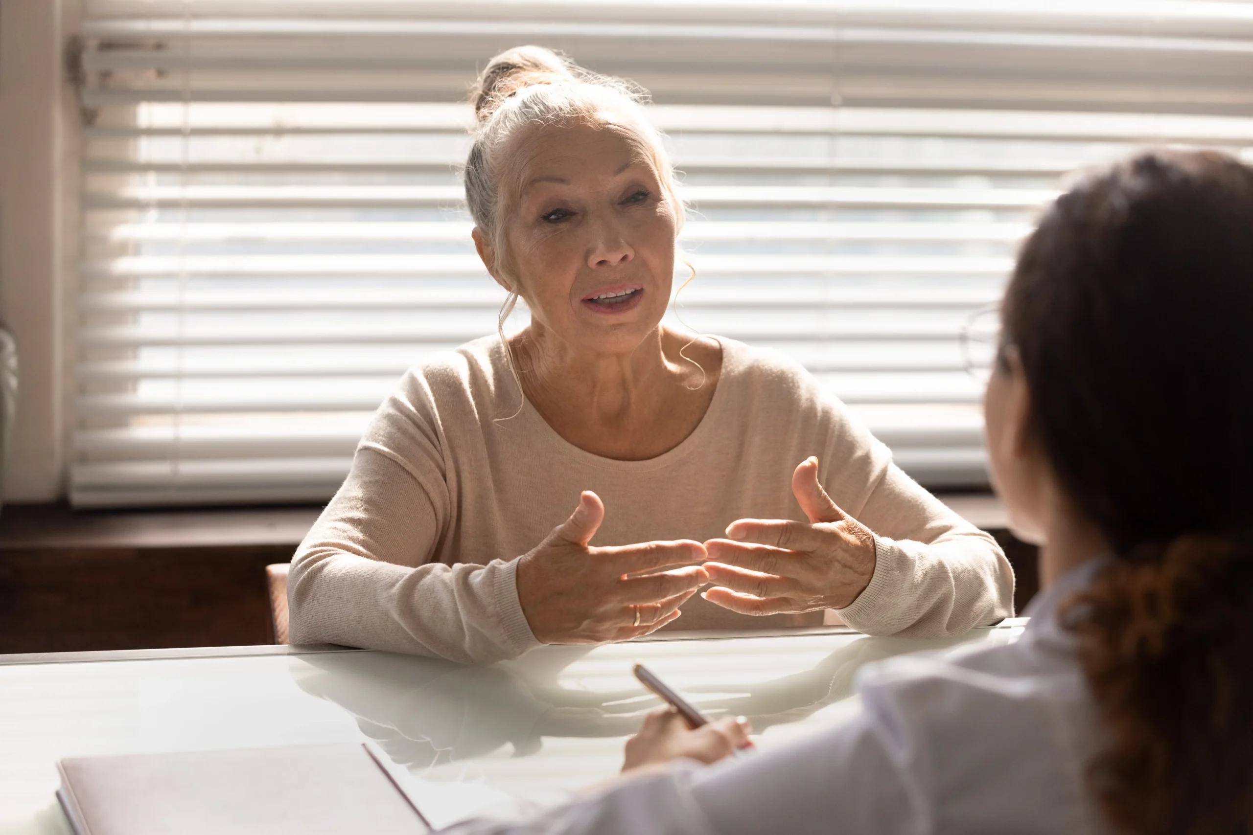 Senior woman with hands gesturing expressively while speaking to therapist during a consultation session in a bright office with natural lighting and window blinds in background
