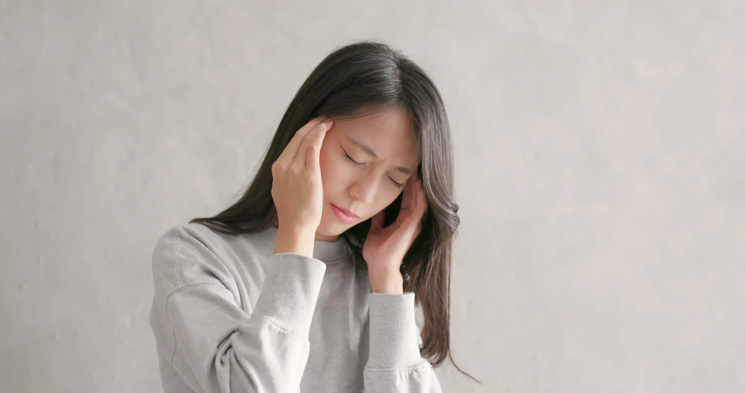 Young woman with long dark hair in gray sweatshirt holding both hands to her head with eyes closed appearing to experience headache or stress against neutral background