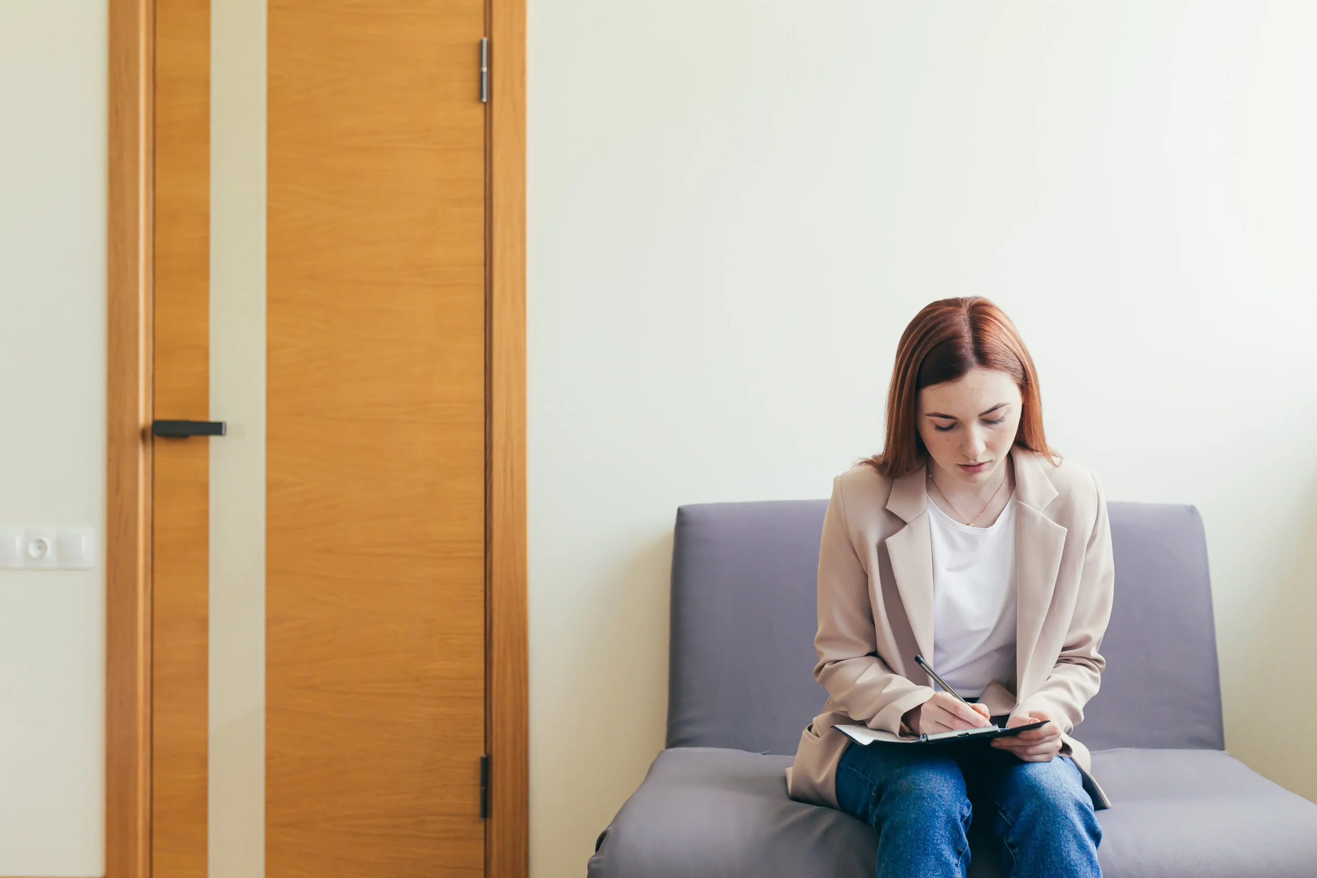 Young woman in beige blazer and jeans sitting on a gray couch writing in a notebook in a minimalist waiting room with white walls and wooden door