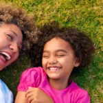 Happy grandmother and young granddaughter with curly hair lying on green grass laughing together, overhead view