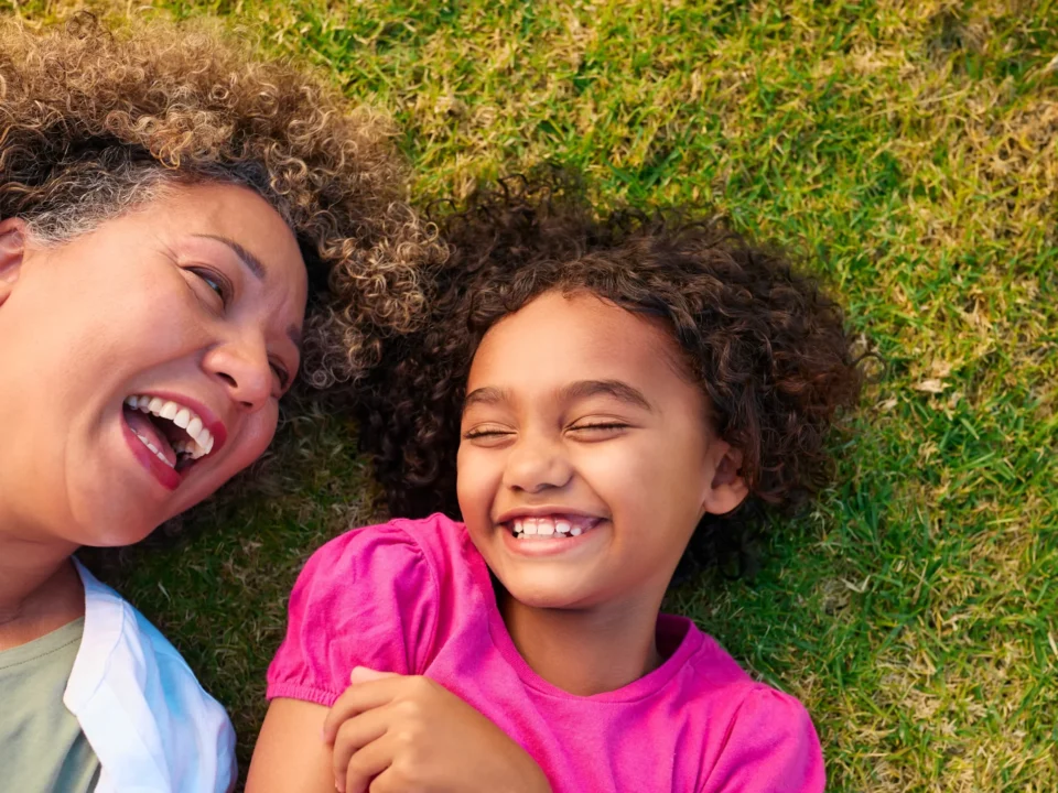 Happy grandmother and young granddaughter with curly hair lying on green grass laughing together, overhead view