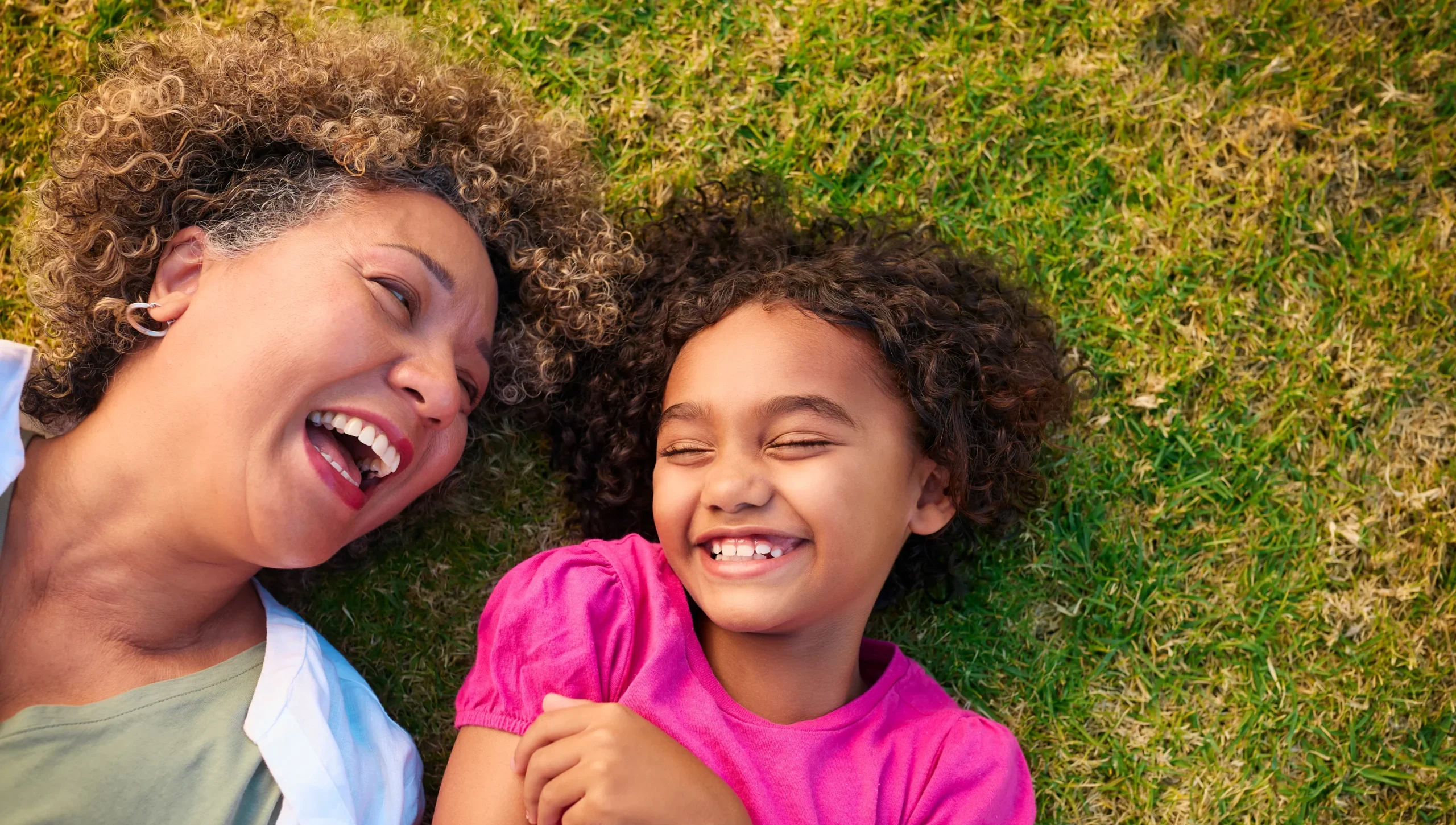 Happy grandmother and young granddaughter with curly hair lying on green grass laughing together, overhead view